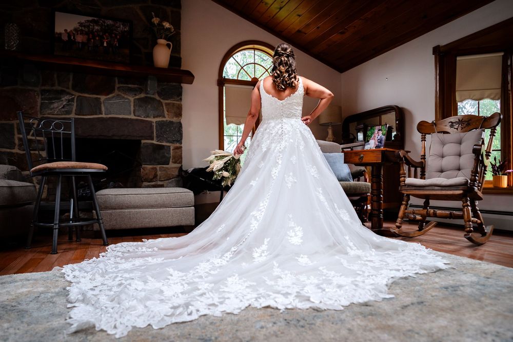 Elegant white wedding dress with intricate lace train displayed in rustic indoor setting with wooden beams and stone walls.