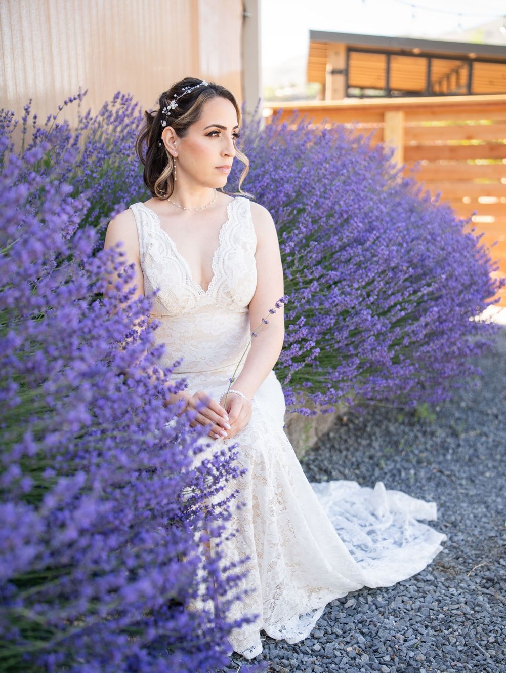 A woman in a lace wedding dress poses beside vibrant purple lavender flowers in a garden setting.