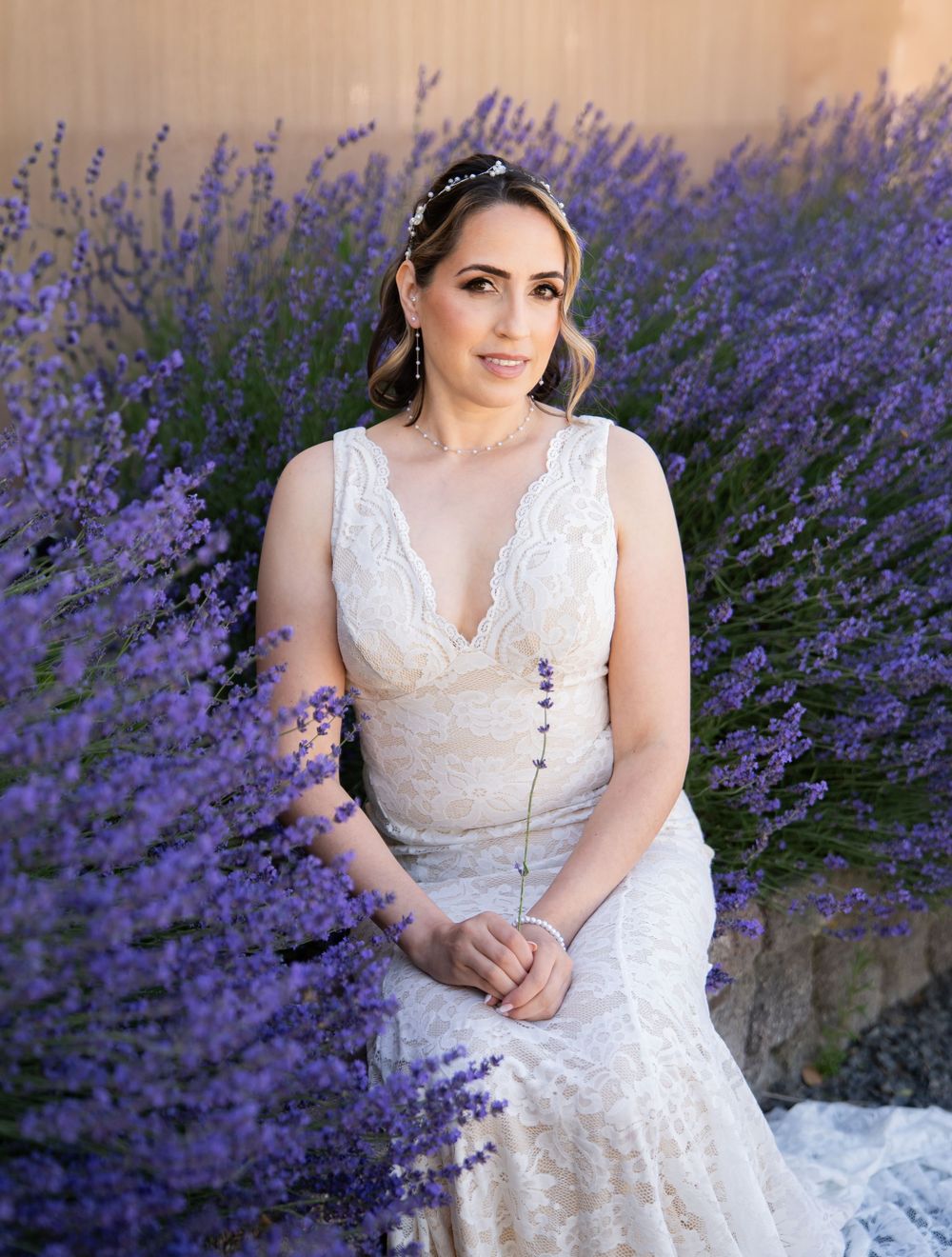 A bride in a white lace dress sits gracefully among blooming purple lavender flowers on a summer day.
