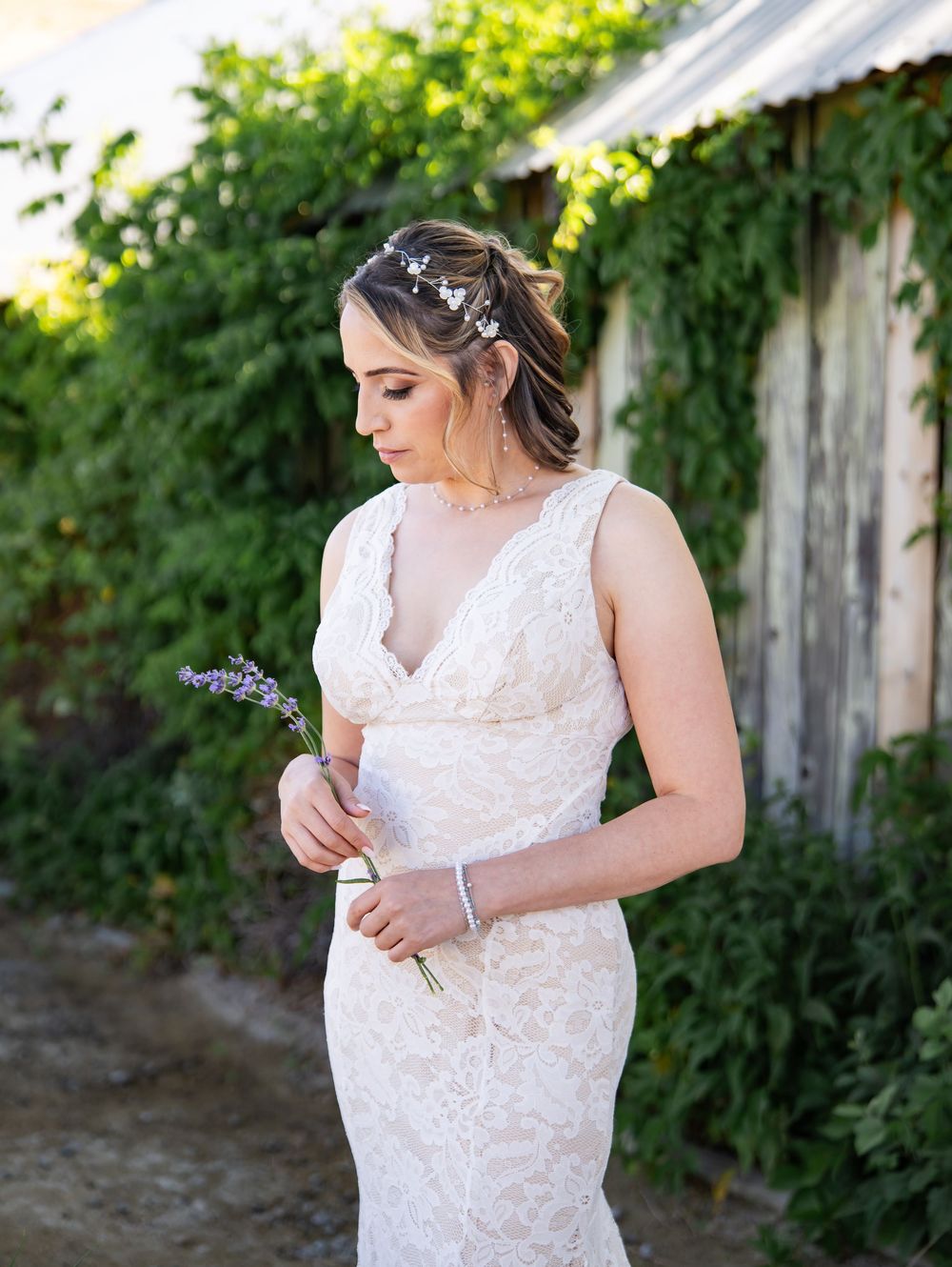 A person in a white lace wedding dress touches delicate details of the gown against a green garden backdrop.