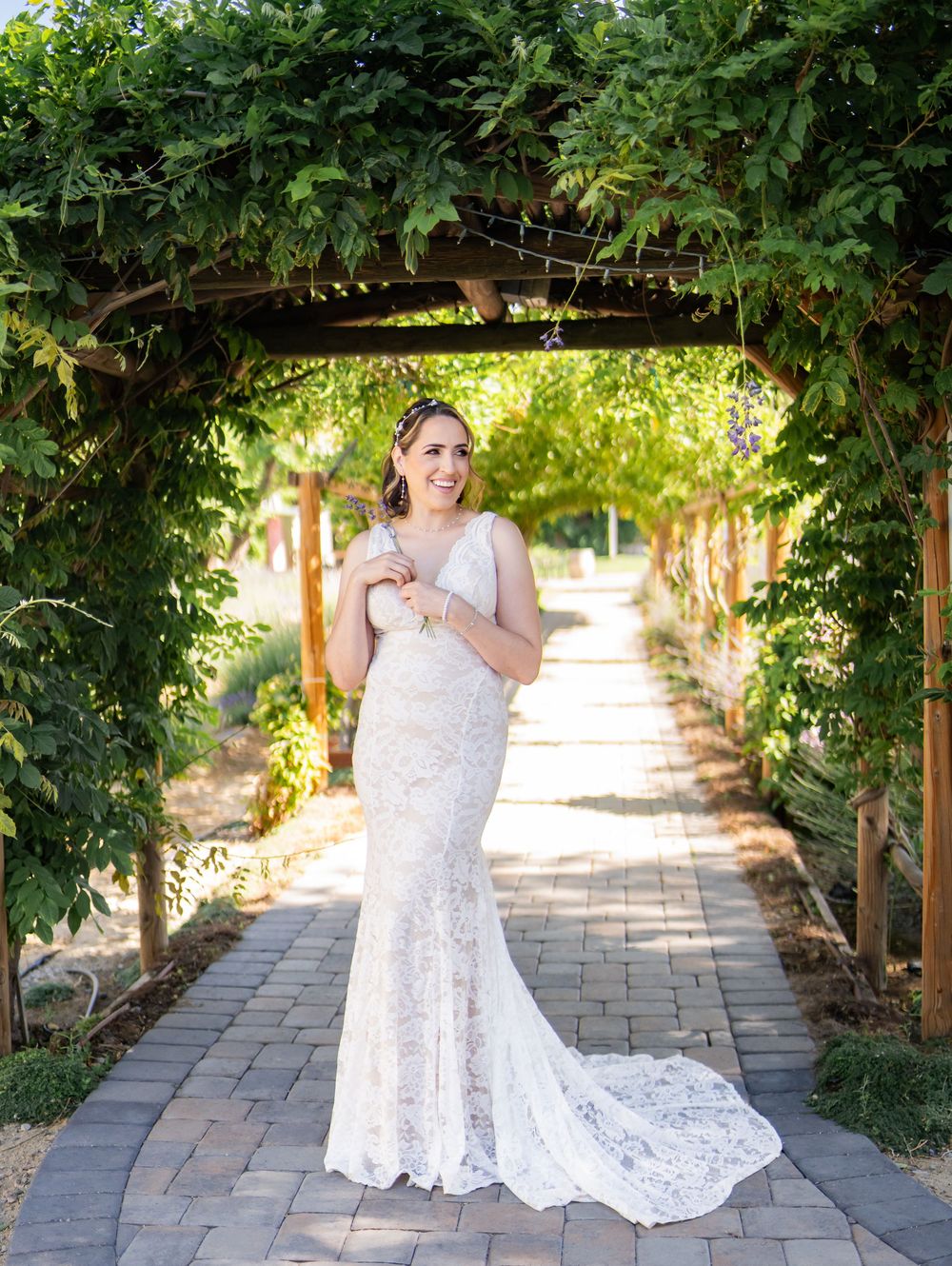 A bride in a lace wedding dress poses gracefully under a vine-covered garden pergola pathway in dappled sunlight.