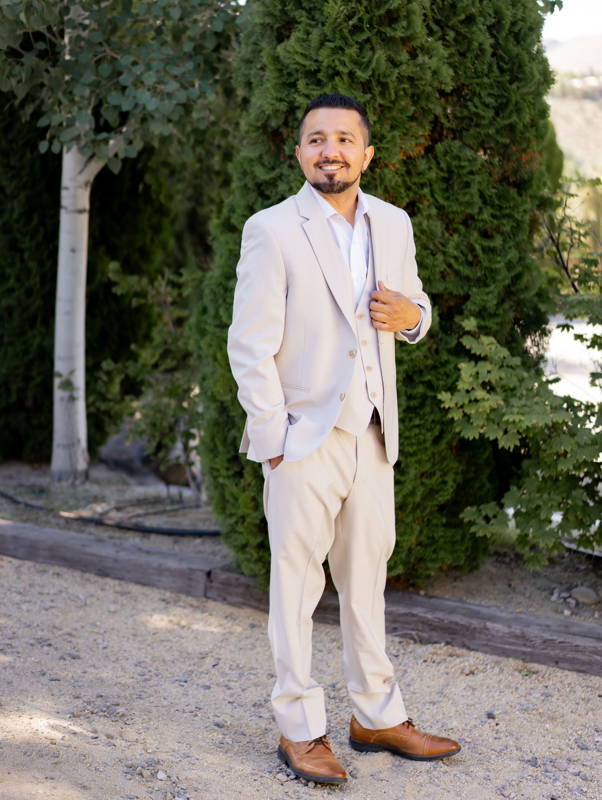 A person in a cream-colored suit poses confidently on a path with green trees in the background.