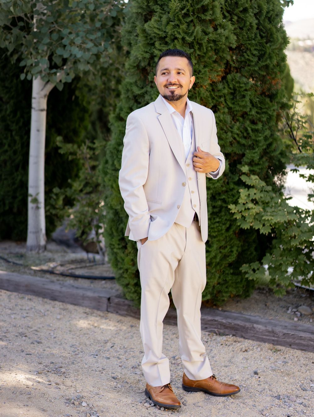 A person in a cream-colored suit poses confidently on a path with green trees in the background.