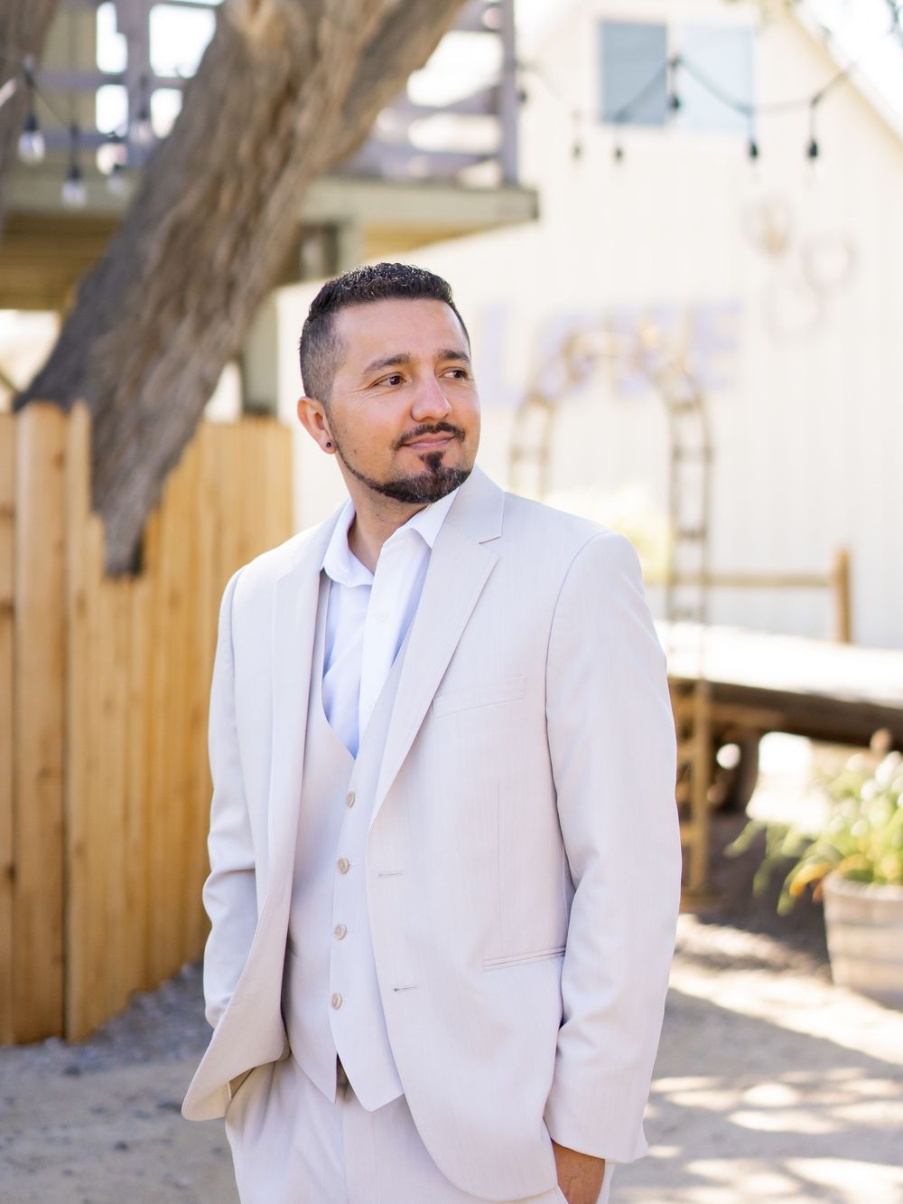 A groom in a light beige suit and white shirt stands outdoors against a rustic wooden backdrop on his wedding day.