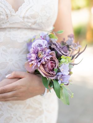 A delicate bouquet of purple and mauve flowers with green foliage held against a white lace wedding dress.