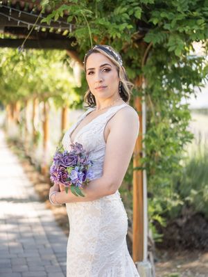 Beautiful bride in lace wedding gown holds purple and blue floral bouquet while posing under garden pergola.