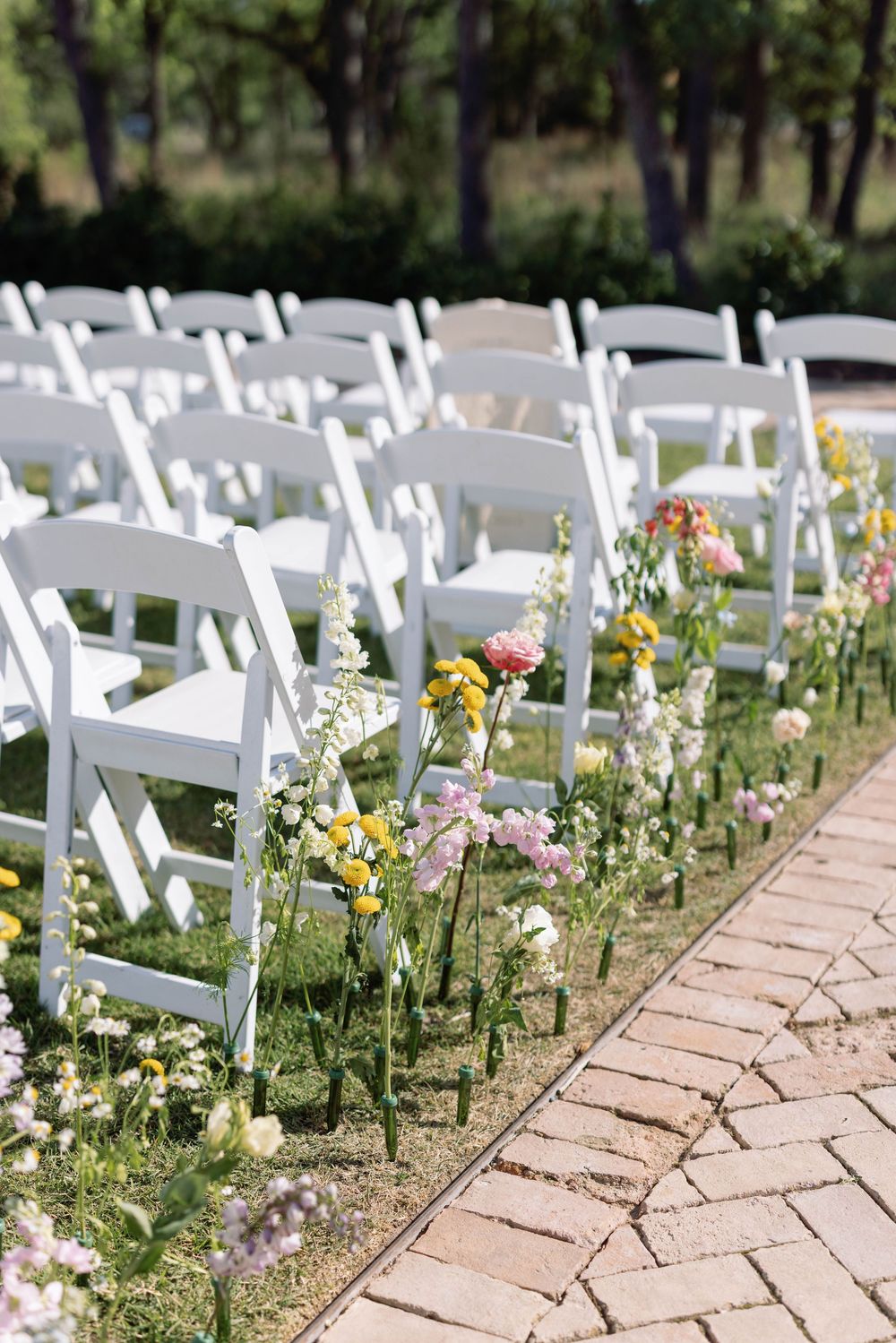 White wedding chairs line a brick pathway with wildflowers and greenery creating a romantic outdoor aisle decoration.