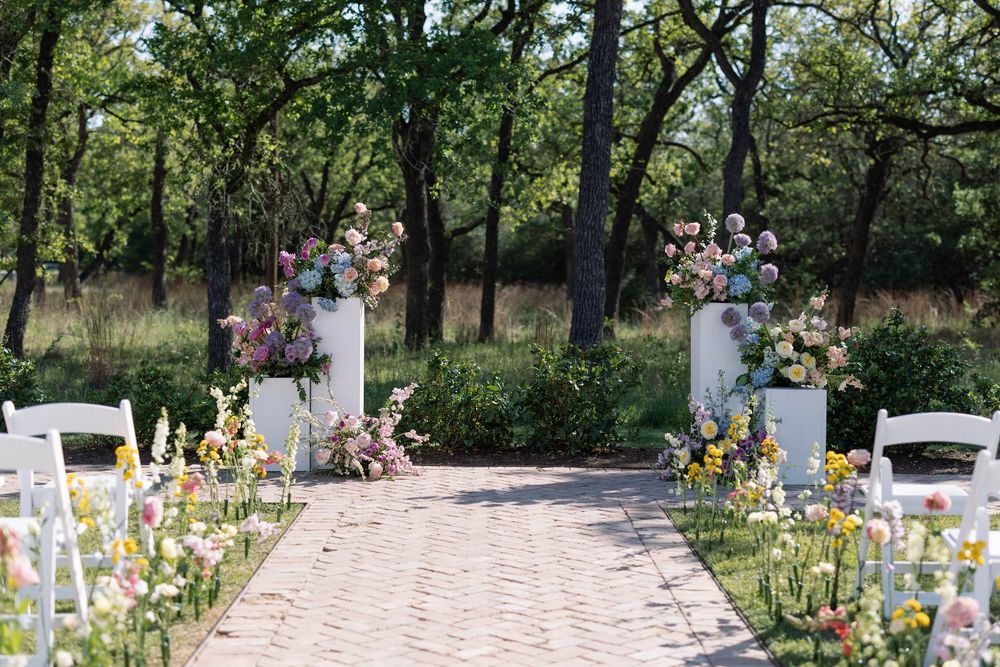 A rustic outdoor wedding ceremony setup with a brick pathway lined with flowers and white chairs in a wooded setting.