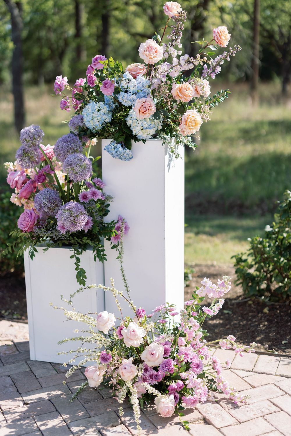 White wooden boxes adorned with lush floral arrangements featuring pink roses, purple blooms and delicate greenery on brick pavers.