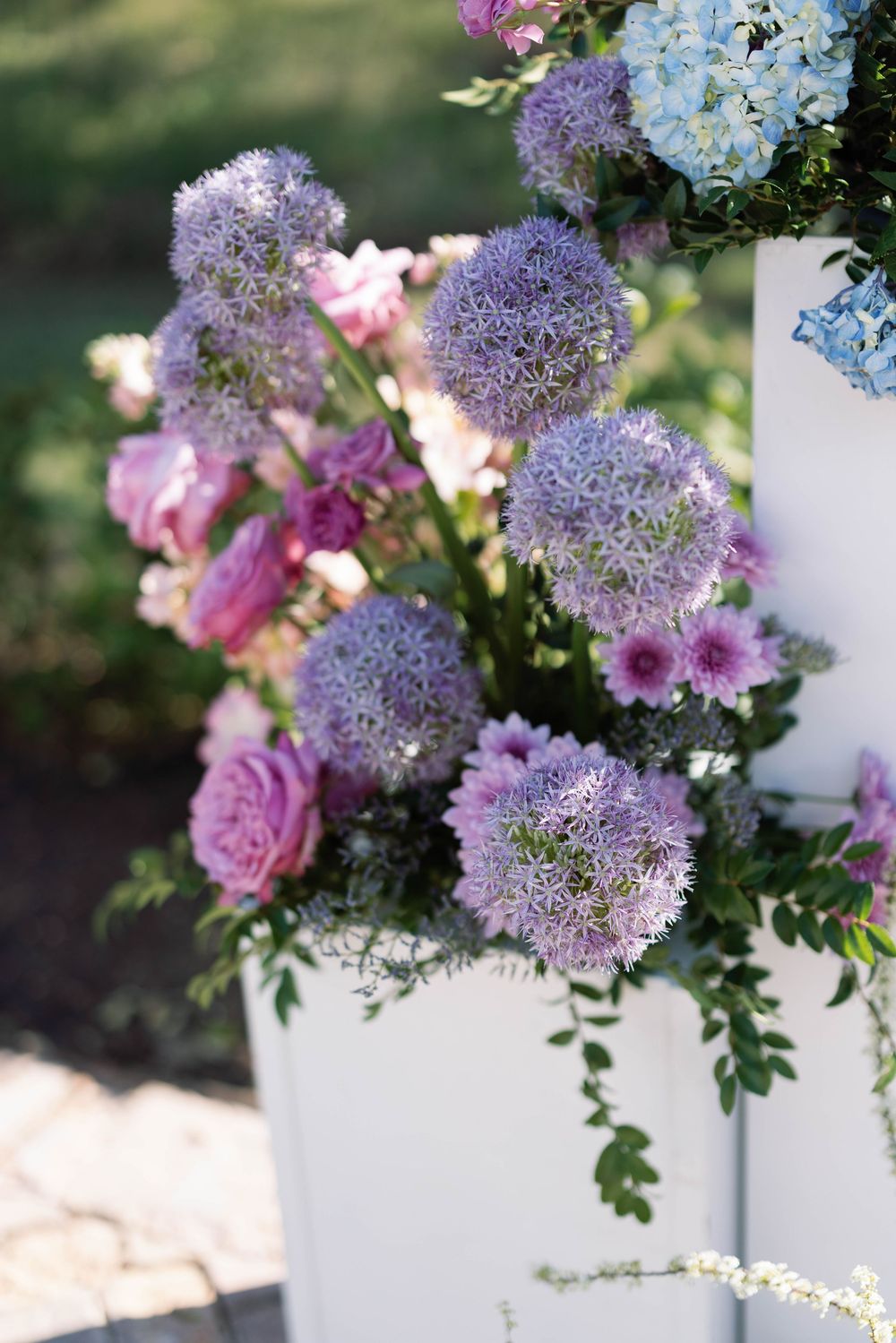 Delicate purple allium flowers and pink roses arranged in a white vase create an elegant floral display outdoors.