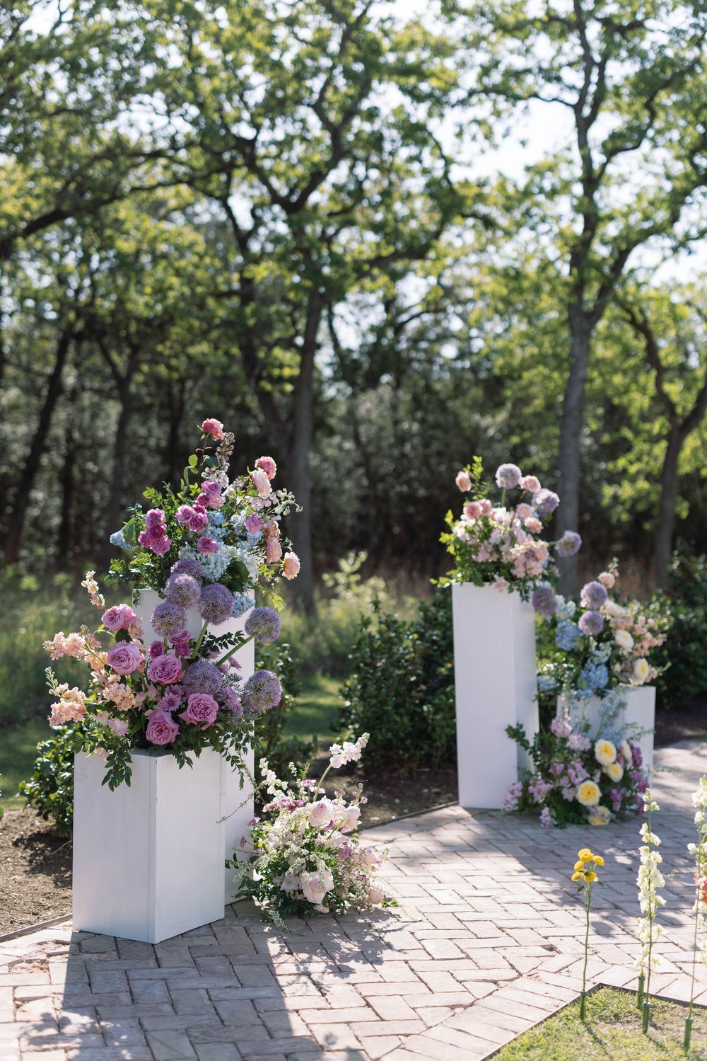 Elegant white pedestals displaying lavish floral arrangements with purple and pink blooms along a stone pathway in a garden setting.