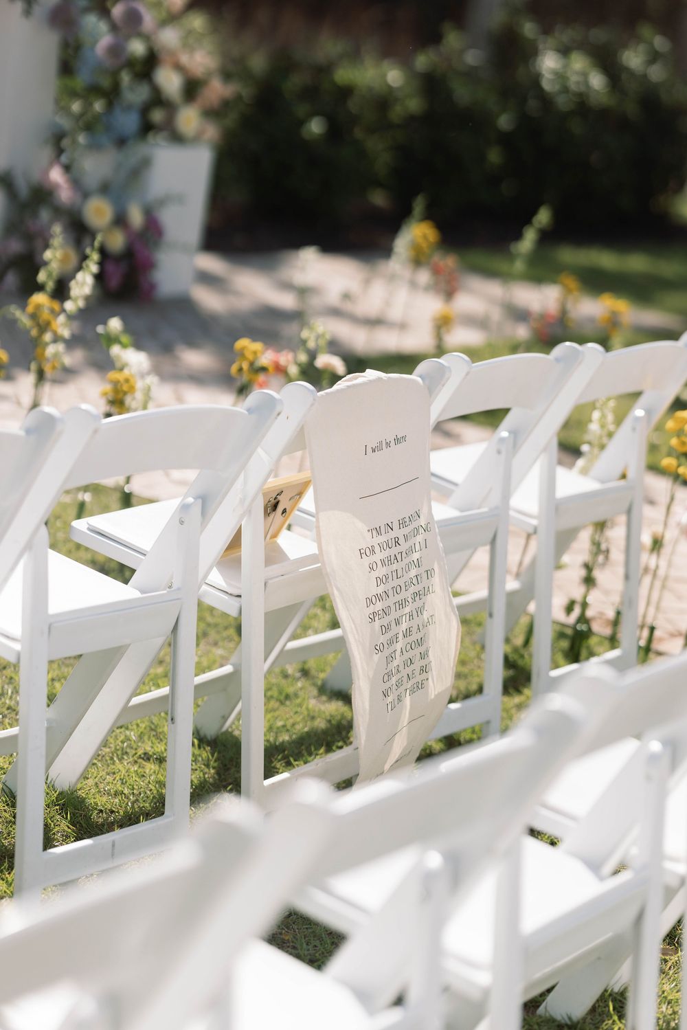 A row of white folding chairs at an outdoor wedding ceremony with a decorative note or program placed on one seat.