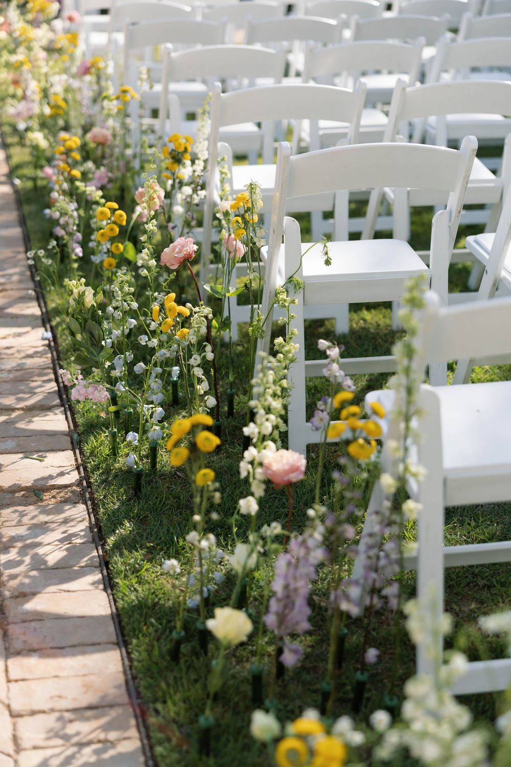 White garden chairs line a wedding ceremony aisle with yellow and pink wildflowers blooming along the pathway.