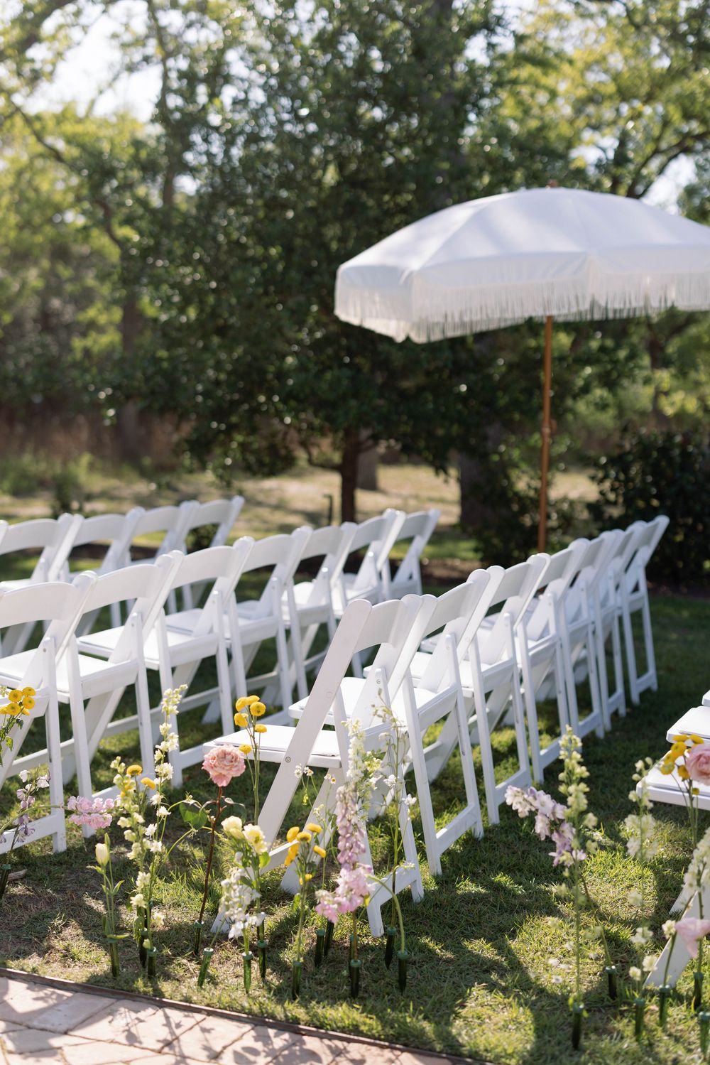 White folding chairs adorned with delicate floral arrangements line up in rows for an outdoor garden wedding ceremony.