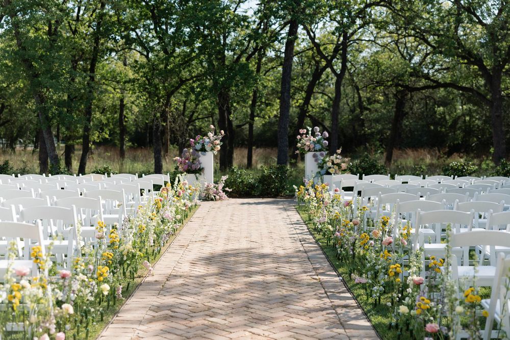 A gravel pathway lined with white crosses and wildflowers stretches through a wooded outdoor memorial.
