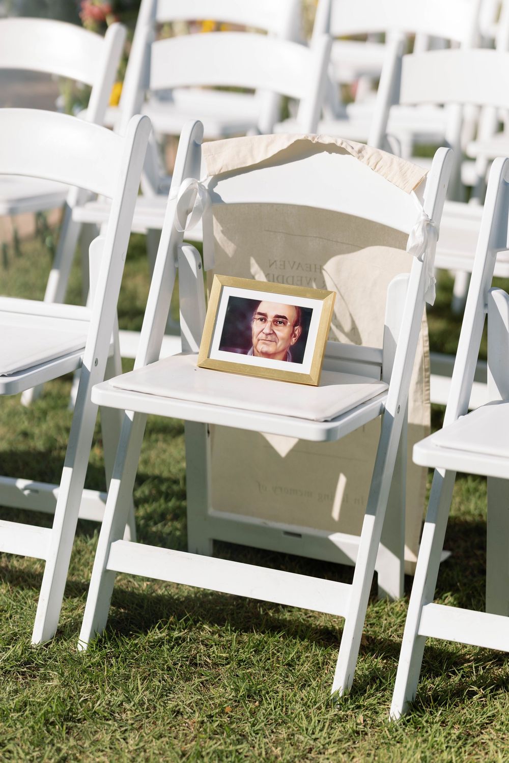 A white folding chair with a framed photo and decorative cloth on the seat for a memorial tribute at a wedding.