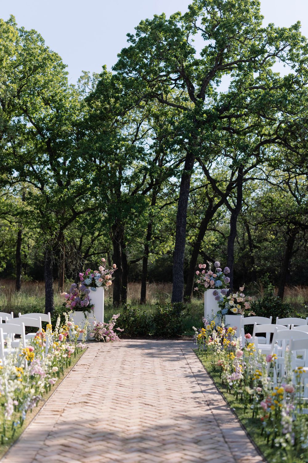 A tree-lined outdoor wedding ceremony setup features a brick aisle bordered by colorful wildflowers and white chairs.