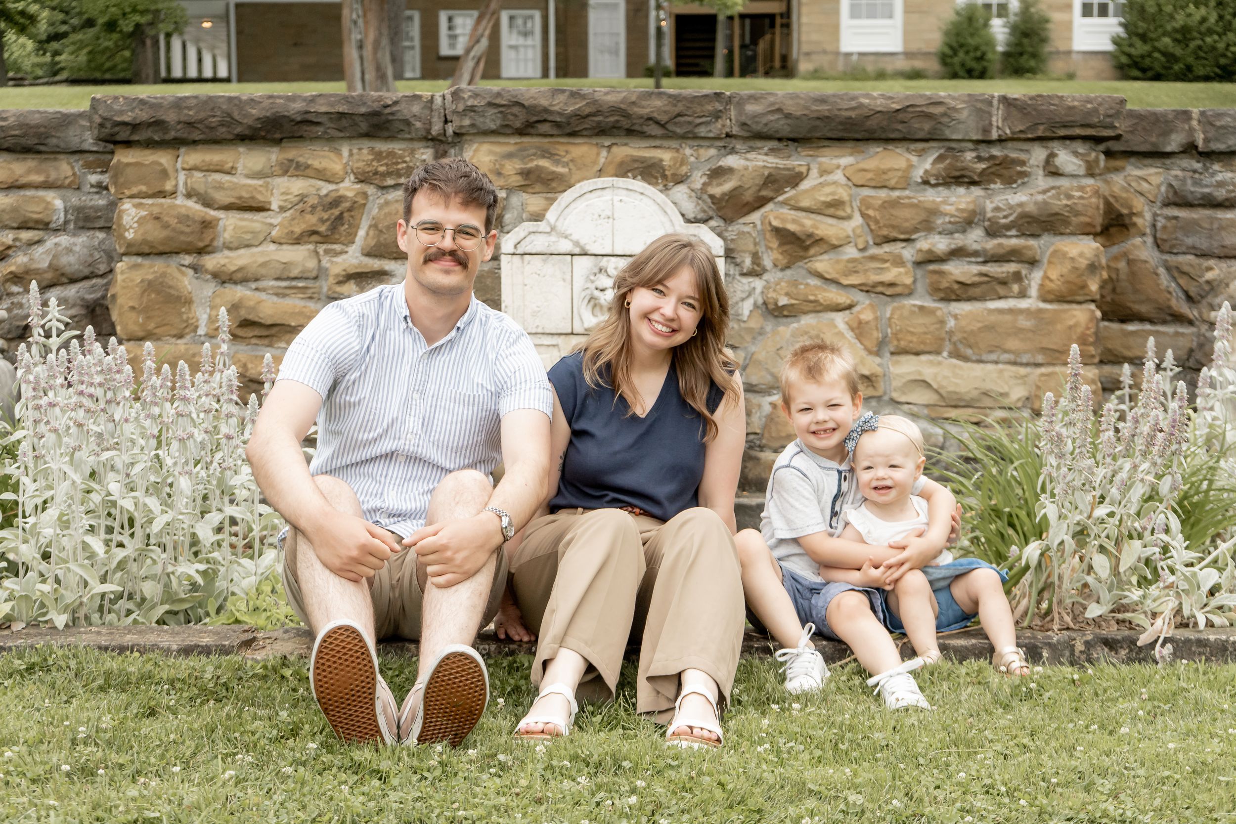 Family of four smiling together outdoors, seated on grass in front of a stone wall with lush greenery.