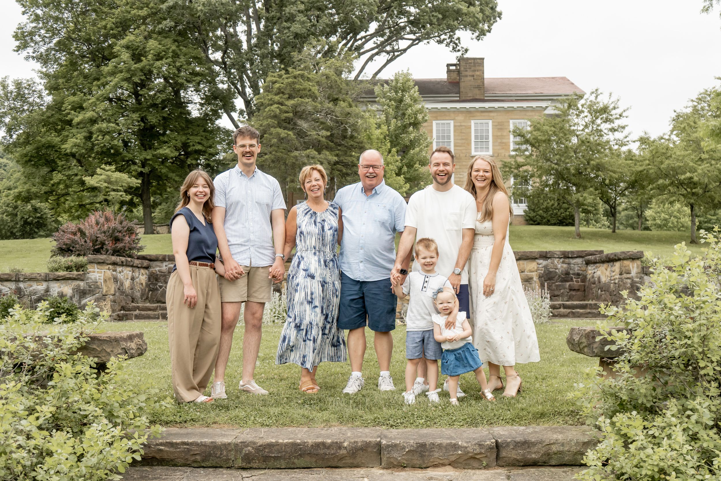 Extended family of seven smiling together outdoors on garden steps with a historic building behind them.