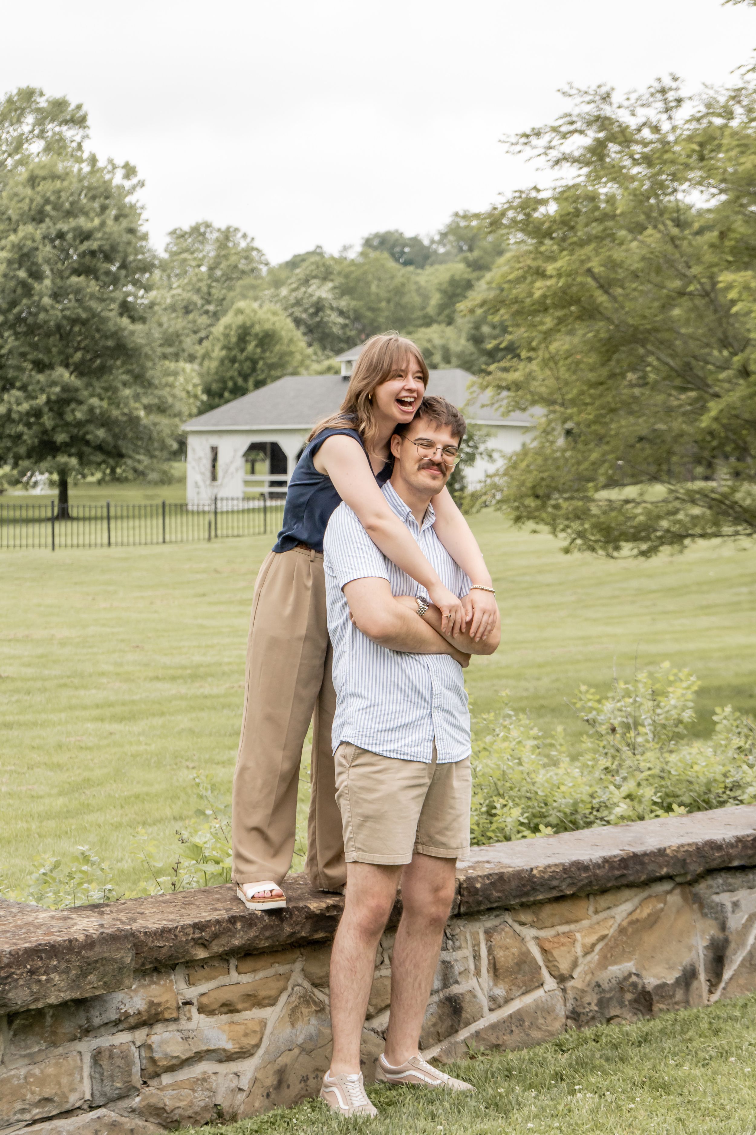 Joyful couple posing on a stone wall outdoors, woman hugging man from behind in a green park.
