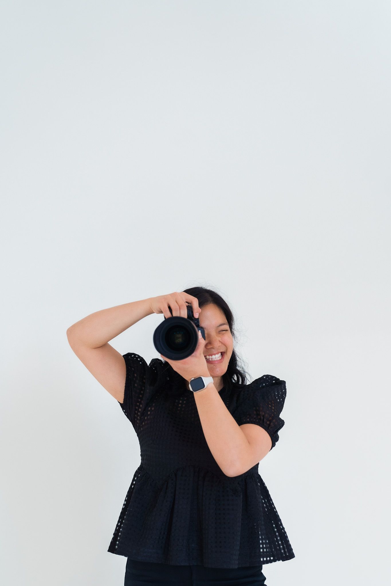 A photographer in a black eyelet lace top holds up a camera against a plain white background.