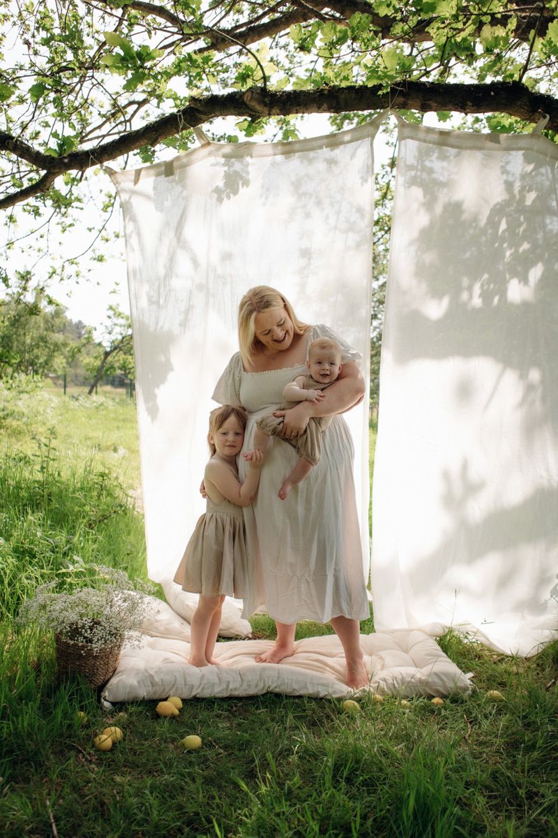 Mother in linen dress holds baby while daughter hugs her outdoors under tree with white fabric backdrop.
