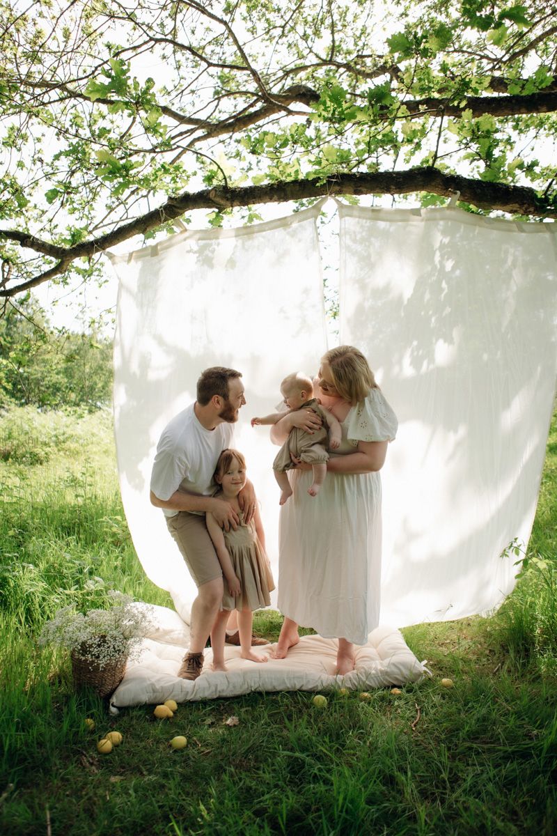 Family of four poses outdoors under a tree branch with white backdrop on lush green grass.