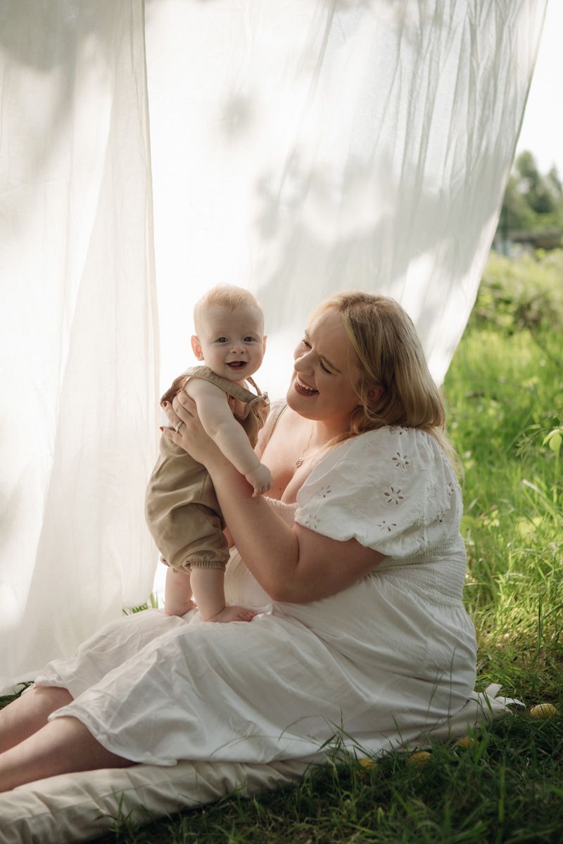 A joyful mother in a white dress sits on grass, lovingly holding her smiling baby outdoors near flowing white curtains.