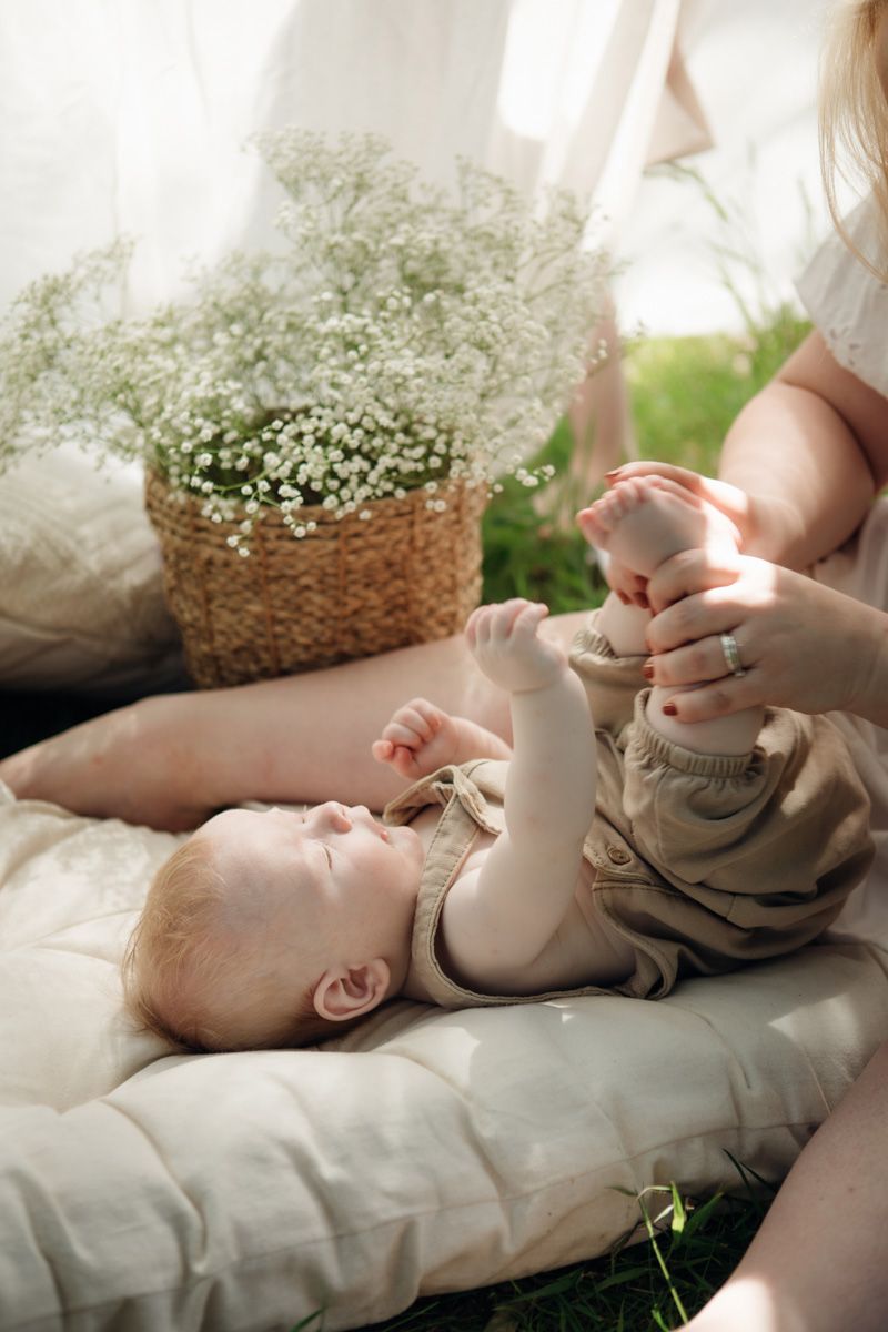 A baby lies on a soft pillow outdoors while a mother gently holds their feet near a wicker basket of white flowers.