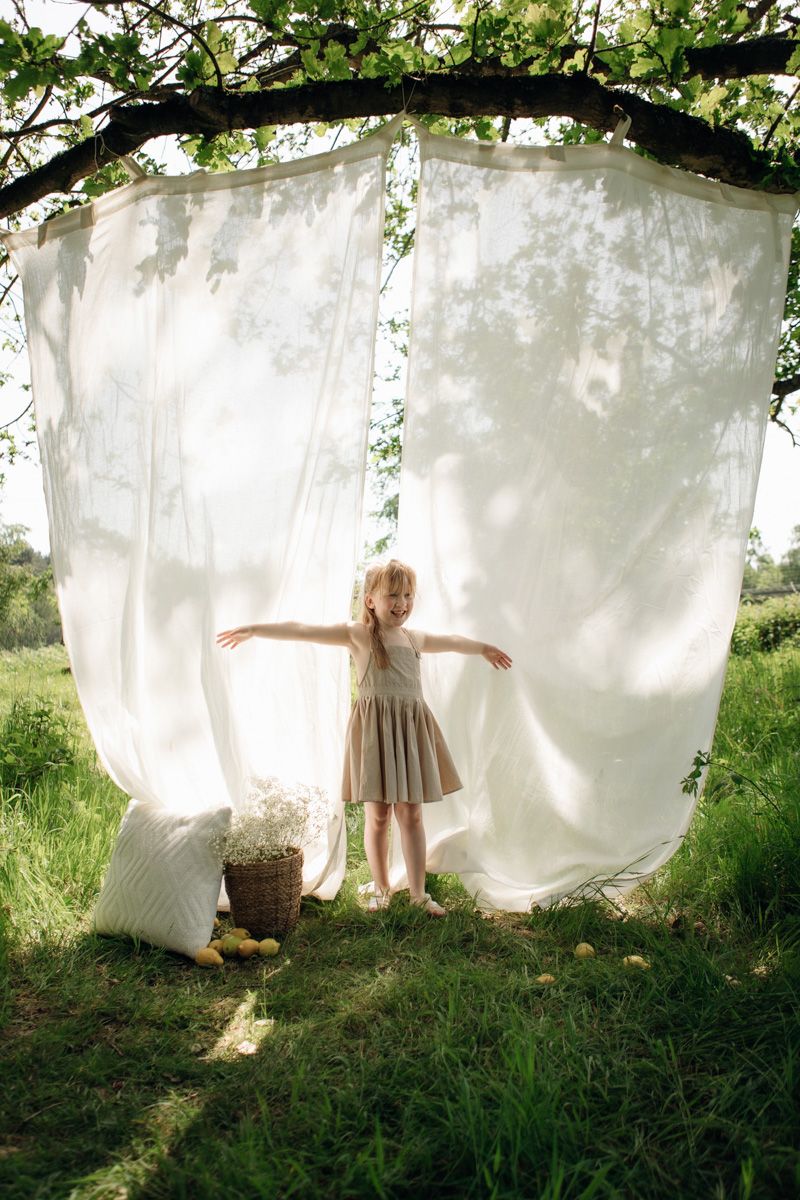 A young girl in a beige dress stands with arms outstretched under white curtains hanging from a tree outdoors.