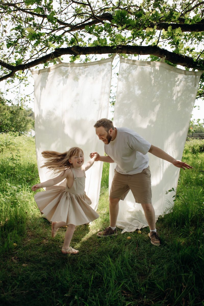 A father and daughter laugh and dance together under white curtains draped from a tree in a green field.