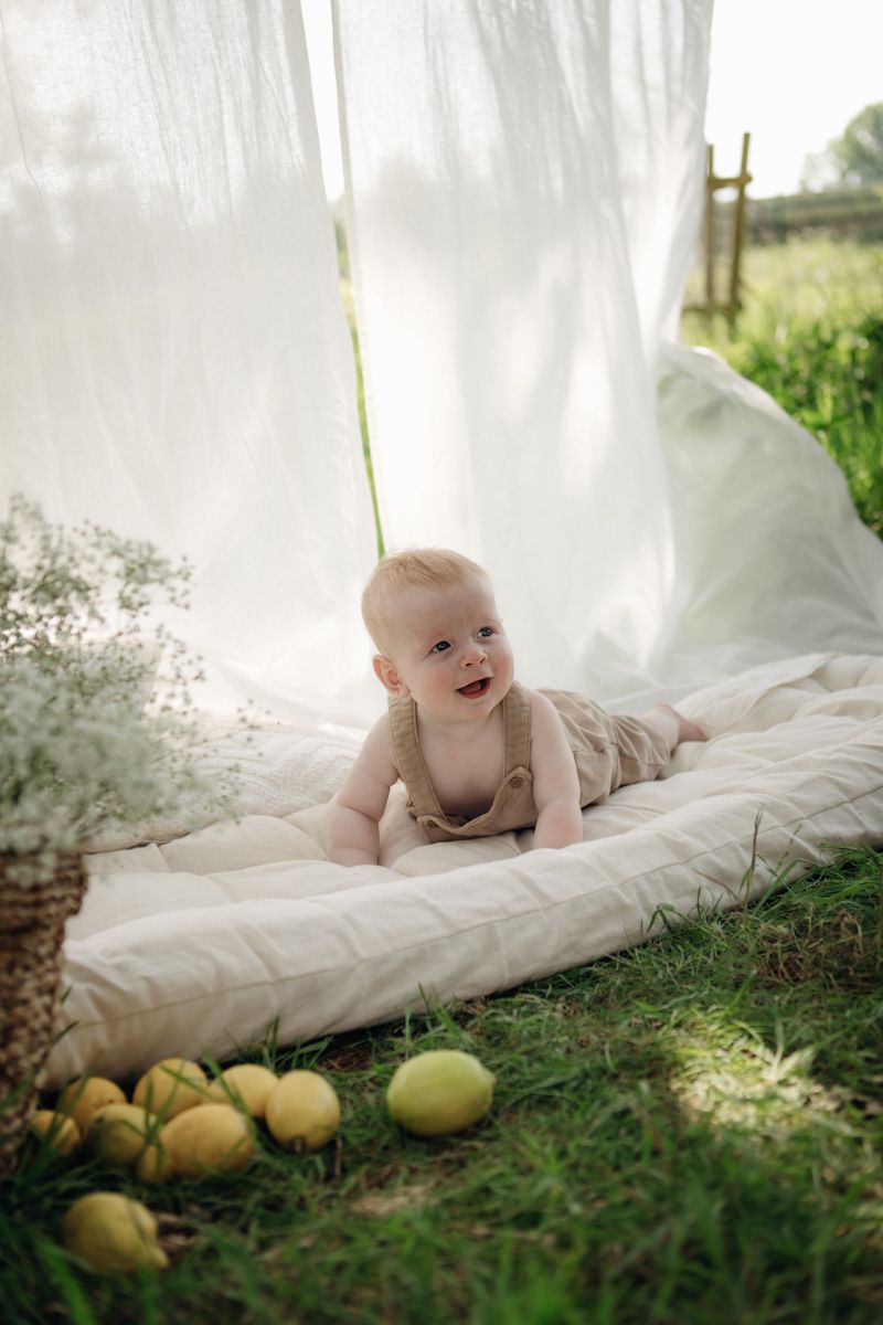 Happy baby in linen overalls lying on a blanket outdoors under white canopy with lemons and flowers nearby.