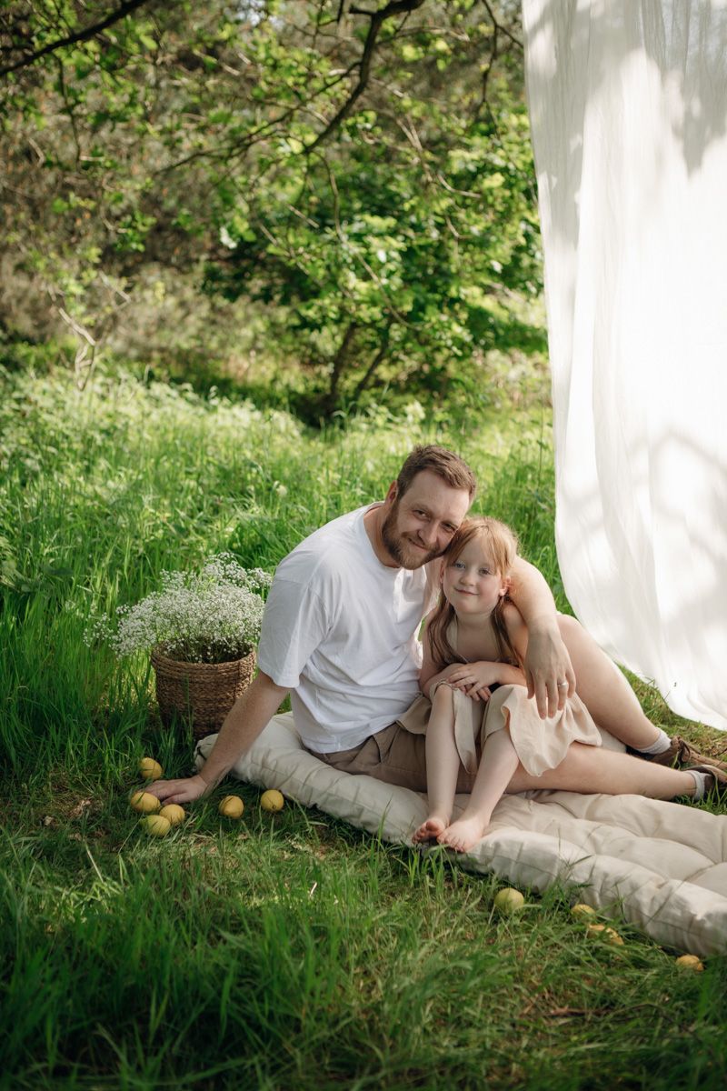 Father and daughter sitting together on a blanket in a lush green garden with apples and white flowers nearby.