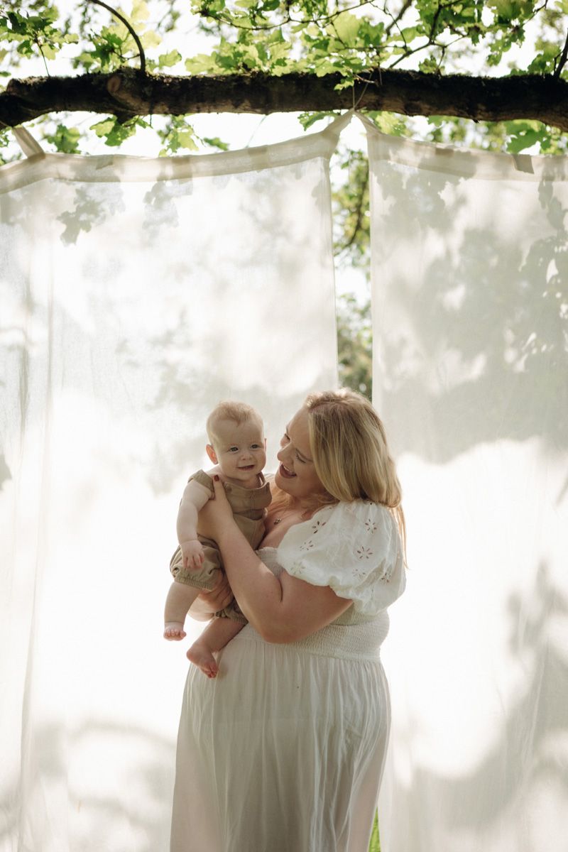 A smiling woman in a white dress holds a happy baby outdoors under sheer white curtains draped from a tree.