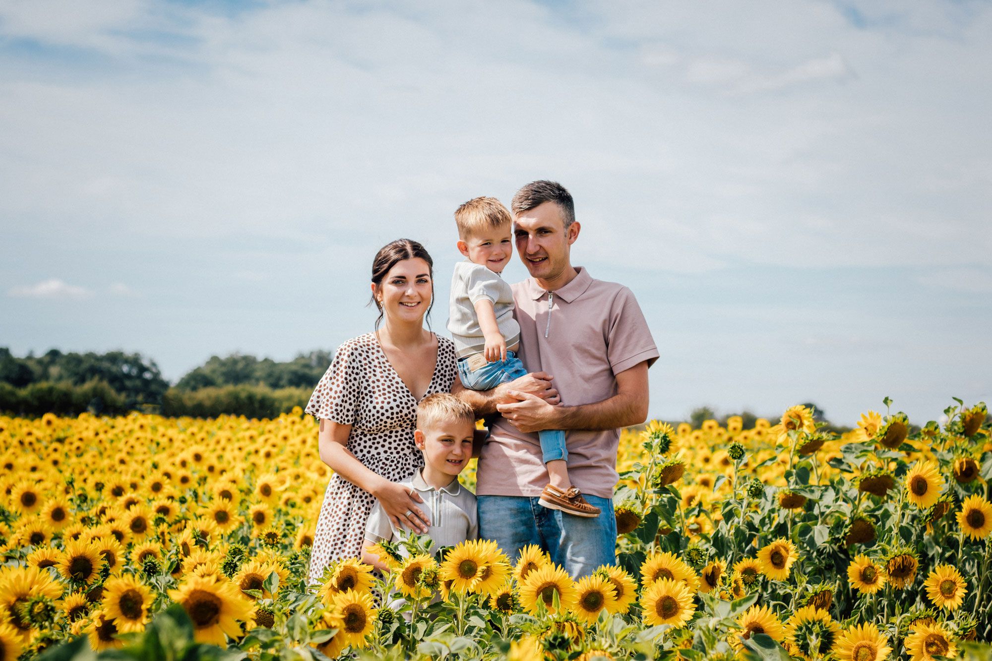 A family stands together in a vibrant field of blooming sunflowers under a partly cloudy summer sky.
