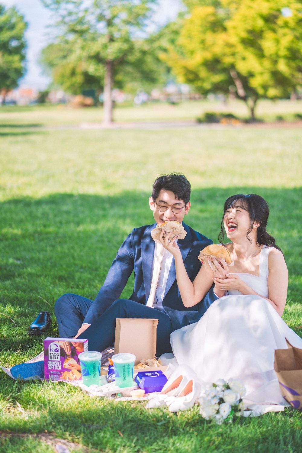 A couple enjoys a casual picnic in a sunny park with snacks and drinks spread out on the grass.