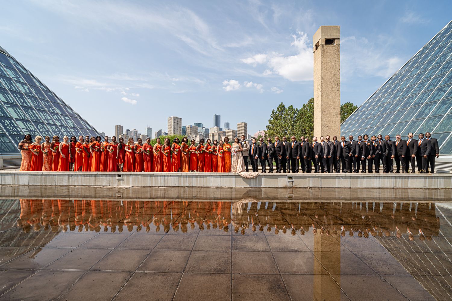 A panoramic collage of people in formal attire standing in rows at a modern architectural venue with glass windows and skylights.