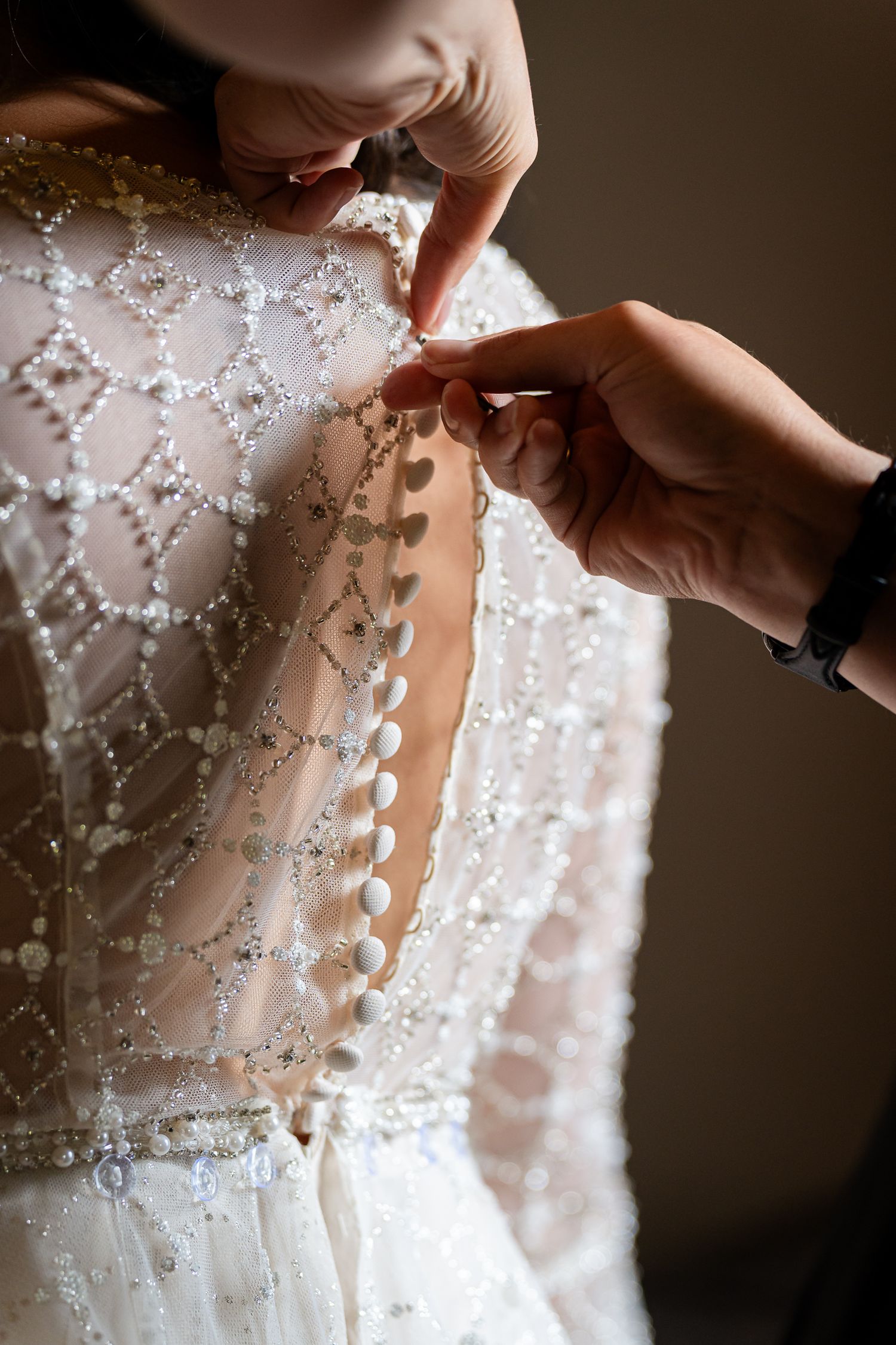 Close-up shots of hands buttoning up a delicate white beaded wedding dress with intricate pearl details.