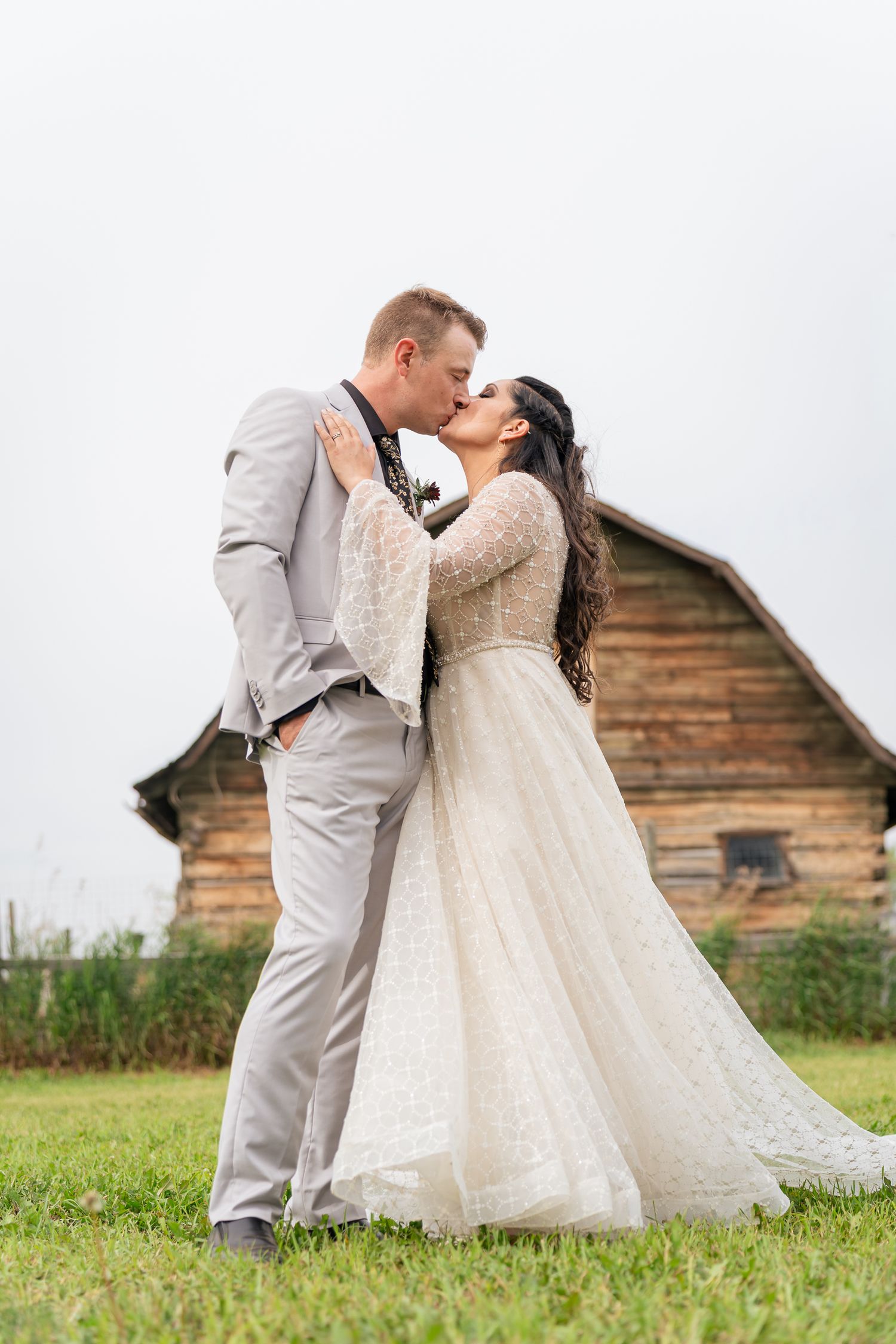 A romantic wedding couple shares a kiss in front of a rustic wooden barn on a grassy field.