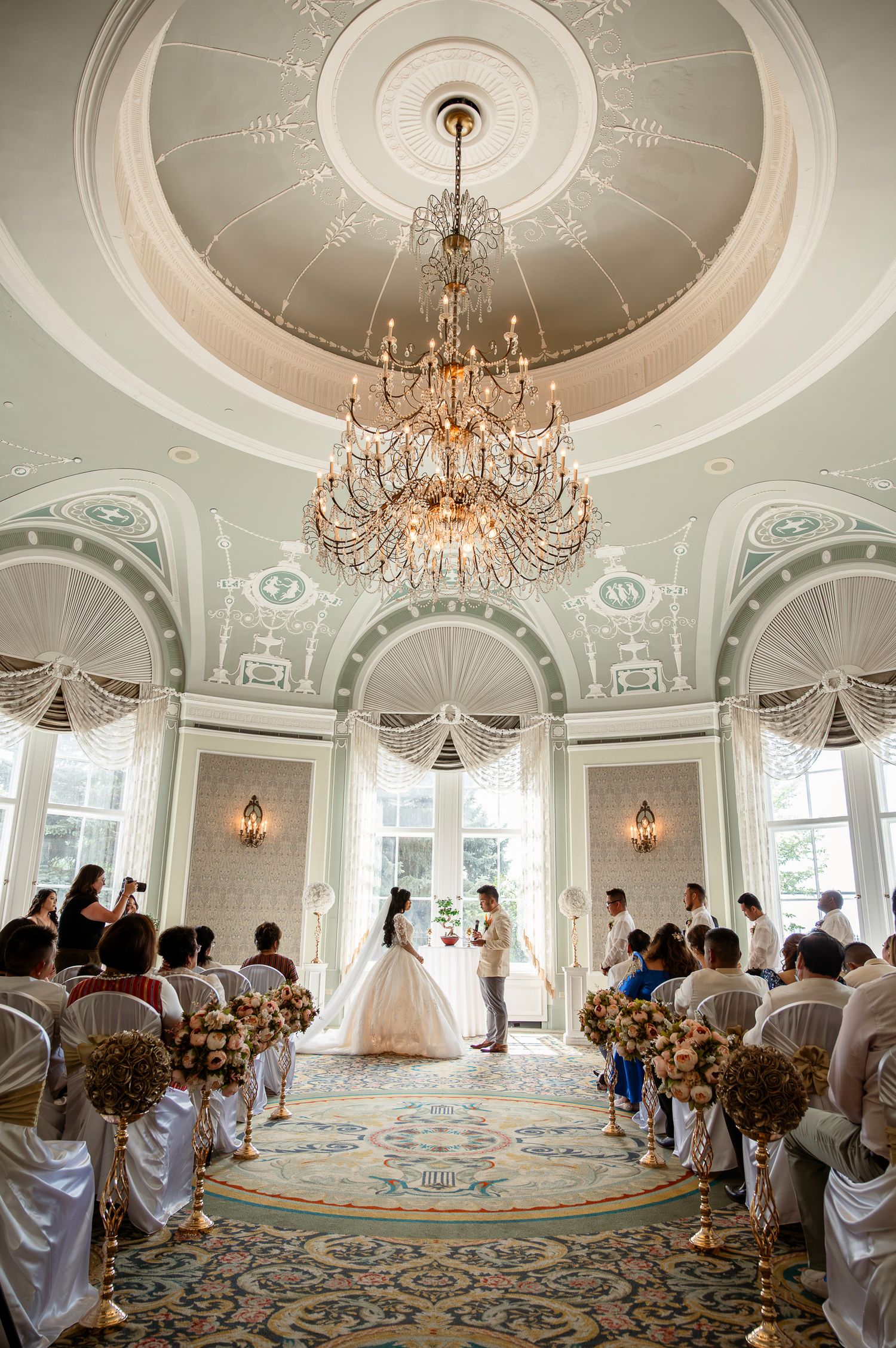 Elegant wedding ceremony in ornate ballroom with crystal chandelier, bride and groom at altar, guests seated in decorated aisles.