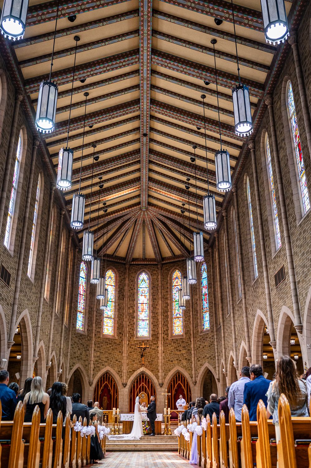A wedding ceremony takes place in a Gothic church with soaring wooden beams, stained glass windows and hanging light fixtures.