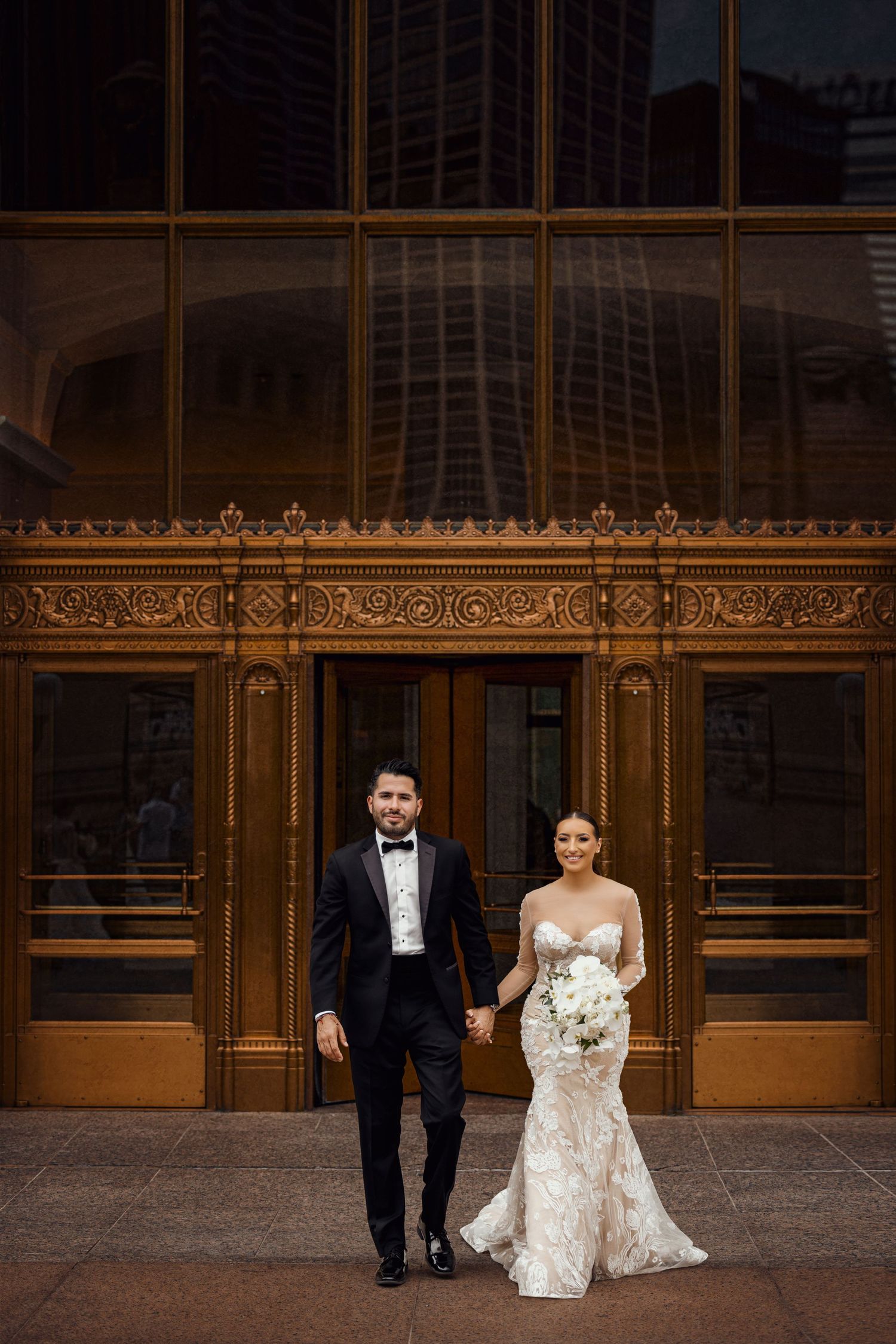 A wedding couple poses in front of ornate golden doors at an elegant building entrance.