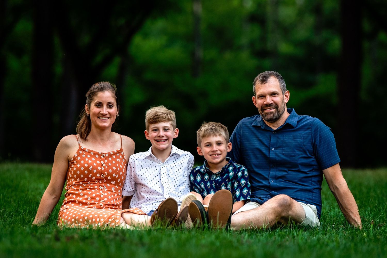 A family of four sits together on green grass with lush trees in the background during a summer day.