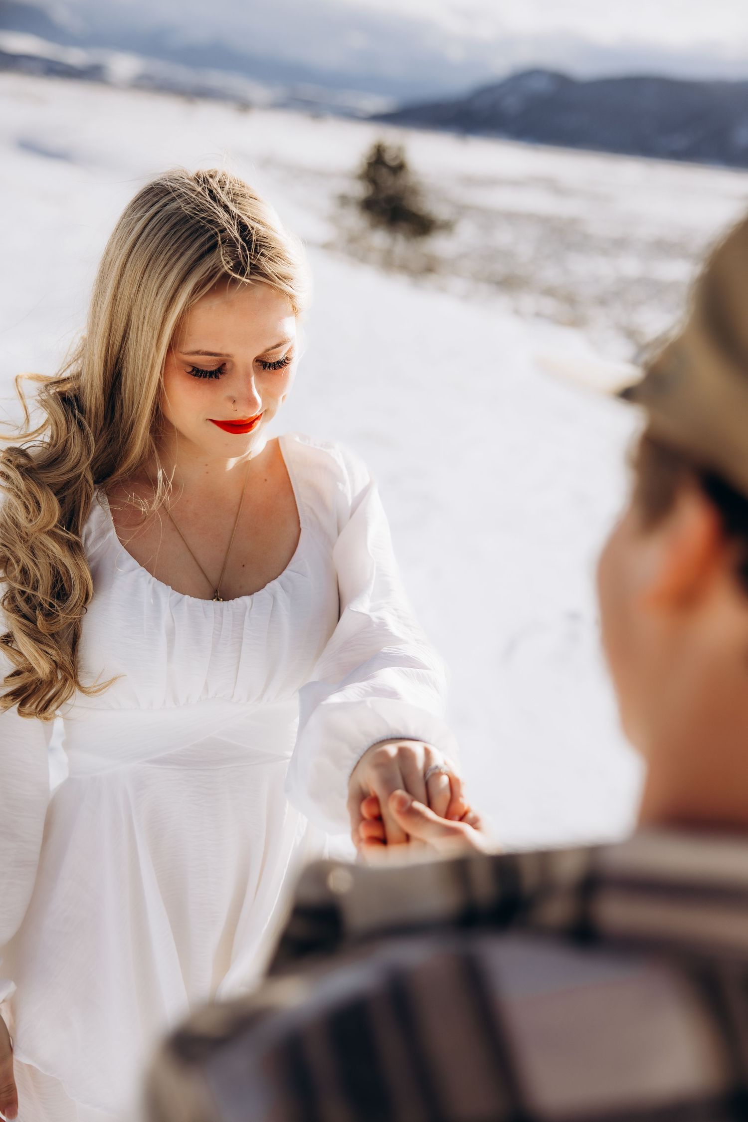 A couple holds hands in a snowy mountain landscape with a white dress and dramatic winter scenery in the background.