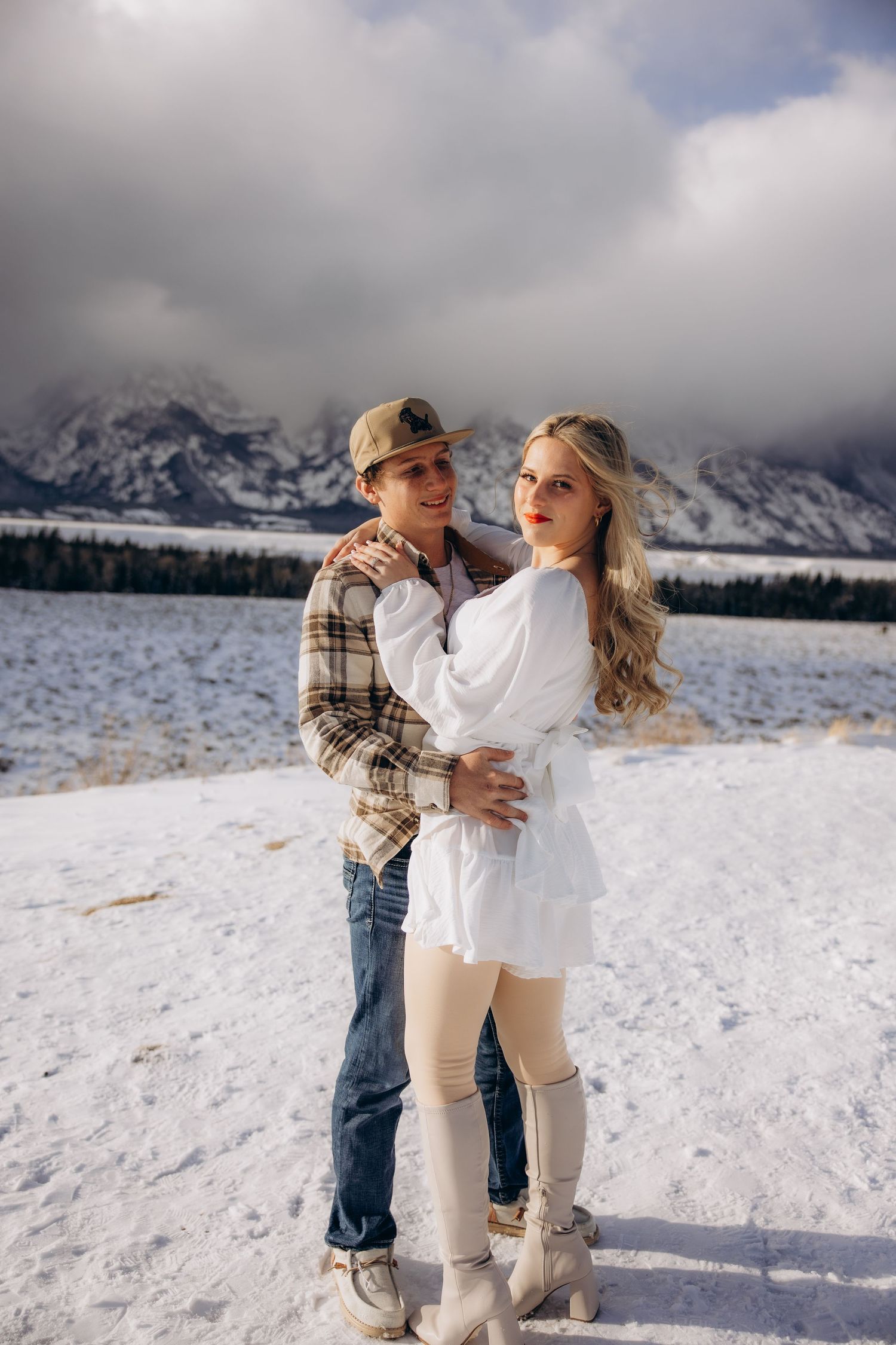 A couple embraces in the snow with dramatic mountain peaks and clouds in the background at Grand Teton National Park.