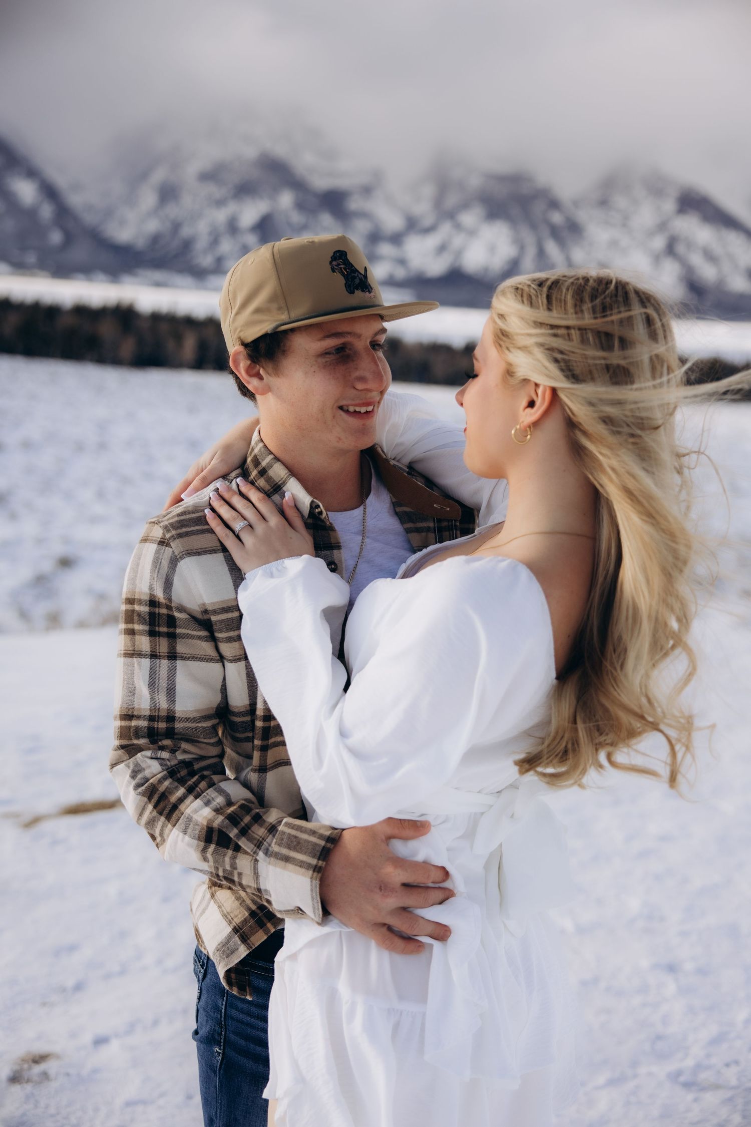 A couple embraces in the snow with majestic snow-capped mountains in the background.