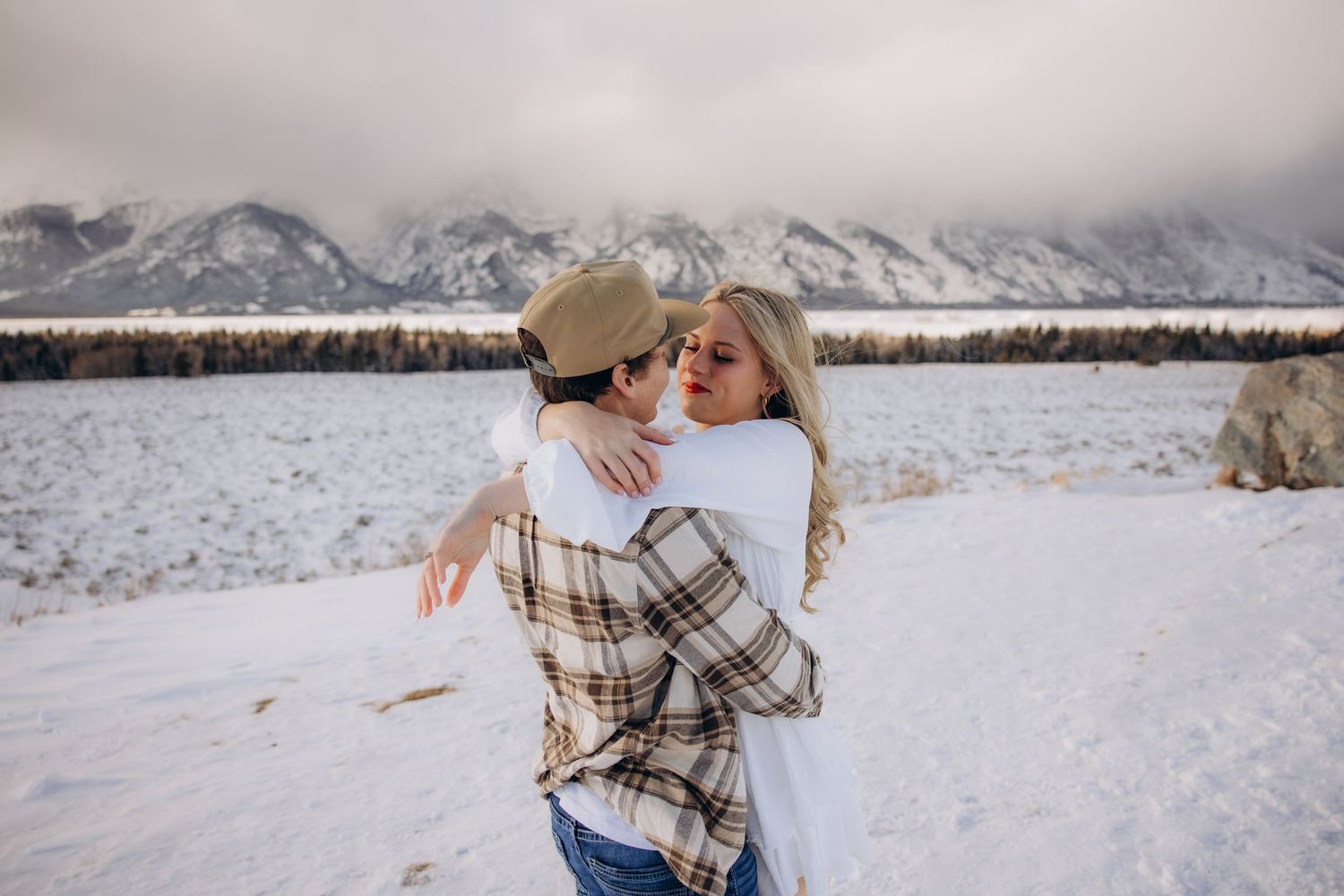 A couple embraces in a snowy field with snow-capped mountains in the background on a winter day.