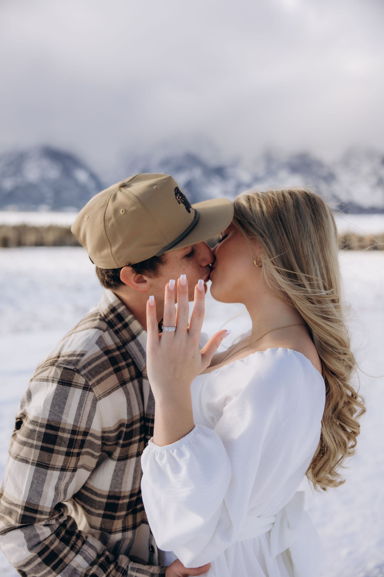 A romantic winter engagement photo showing a couple kissing in front of snow-capped mountains in a snowy field.