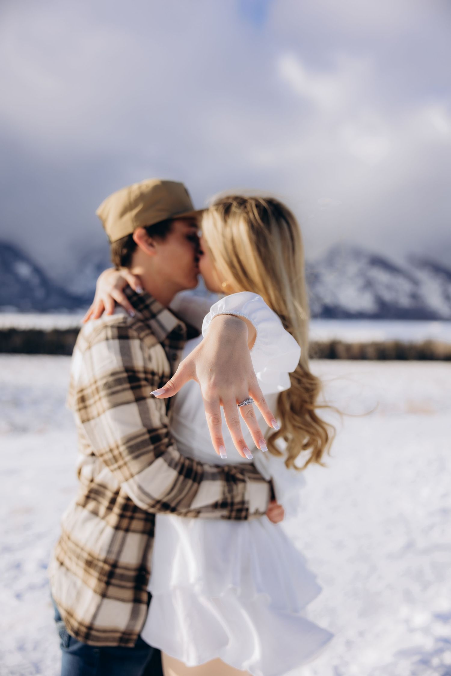 A romantic winter engagement photo shows a couple embracing in the snow with snowy mountains in the background.