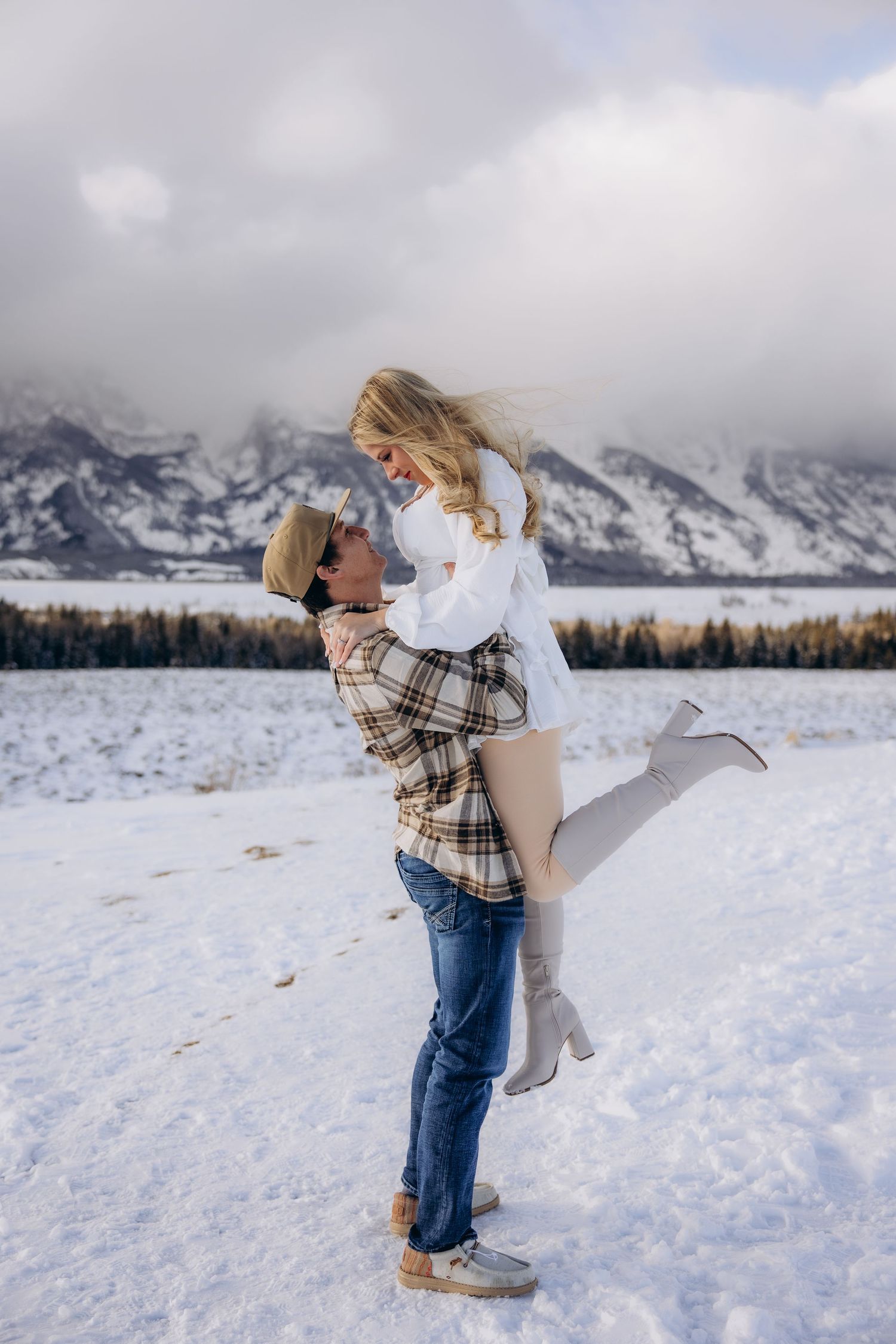 A romantic winter engagement photo shows a couple embracing in front of snow-covered mountains in Grand Teton National Park.